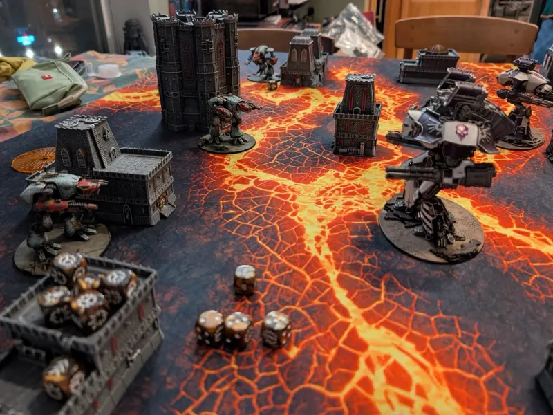 Adeptus Titanicus battlefield on a glowing lava mat: white and purple titans facing weathered green titans among gothic towers.