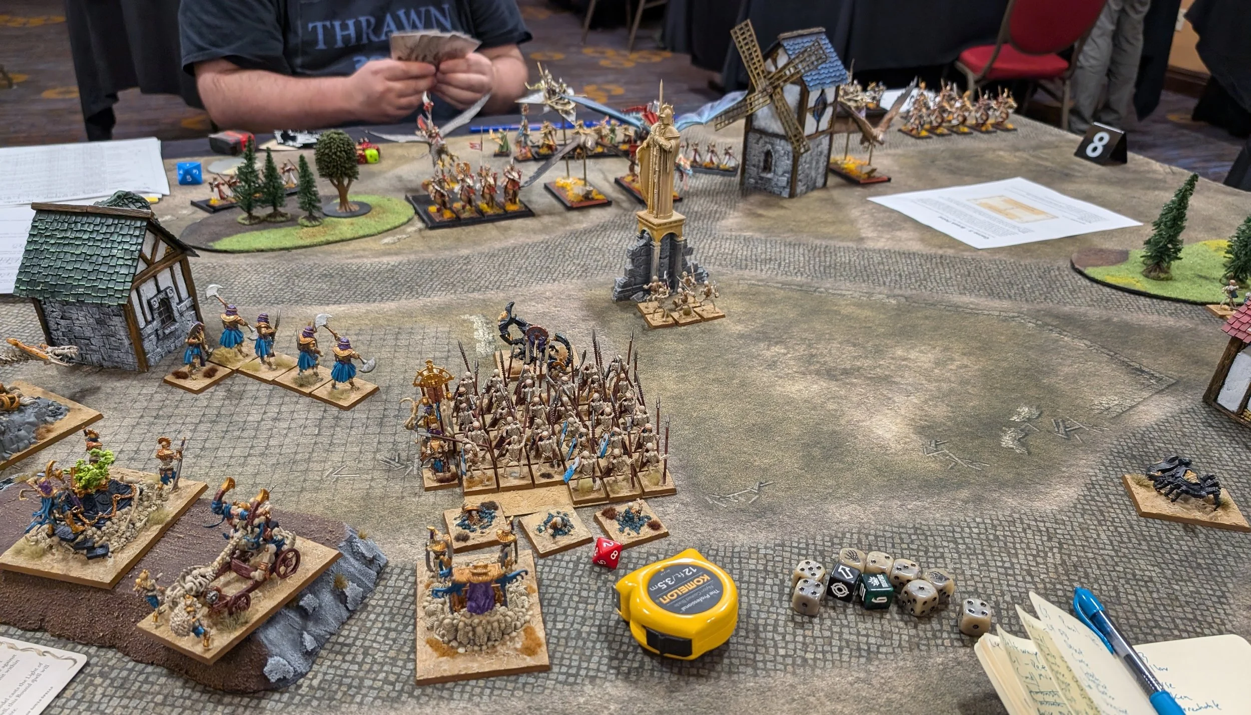 A cobbled village table: a large skeleton spear block and Ushabti advance past a shrine, with High Elf cavalry and a windmill on the far side.