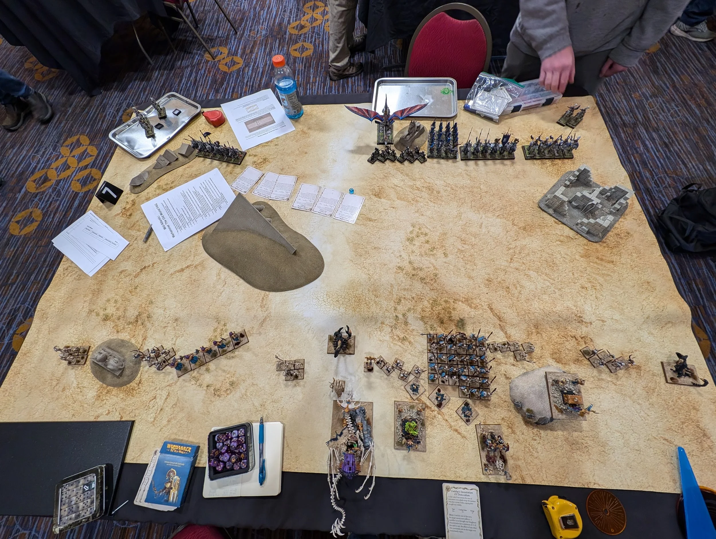 Overhead shot of a desert gaming mat at deployment: Tomb Kings units strung along the near edge, blue-clad High Elf regiments and a dragon along the far edge.