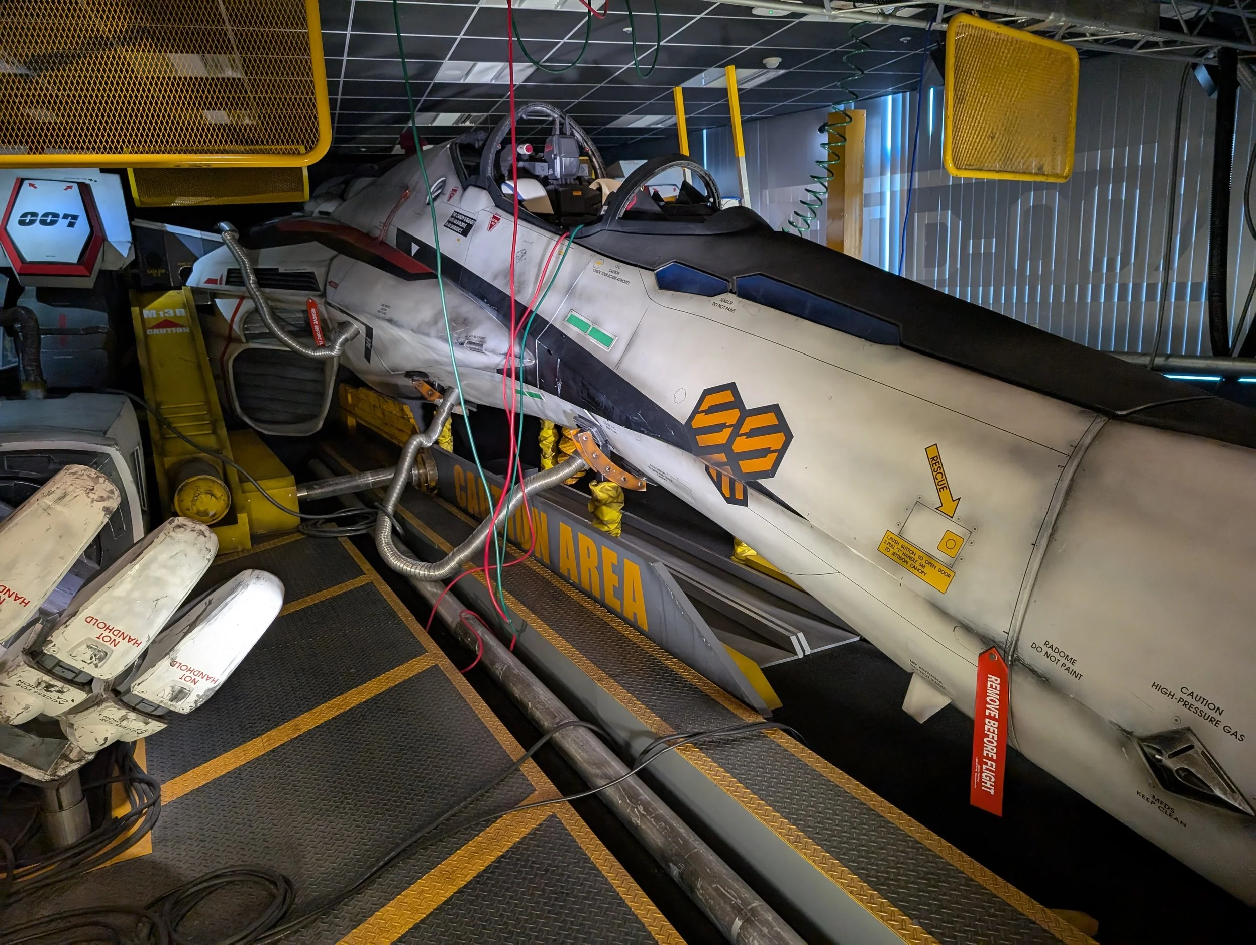 Full-size Valkyrie VF-25F fighter mock-up in a hangar-styled exhibit, hoses and cables running to it across a yellow-striped "Caution Area" floor.