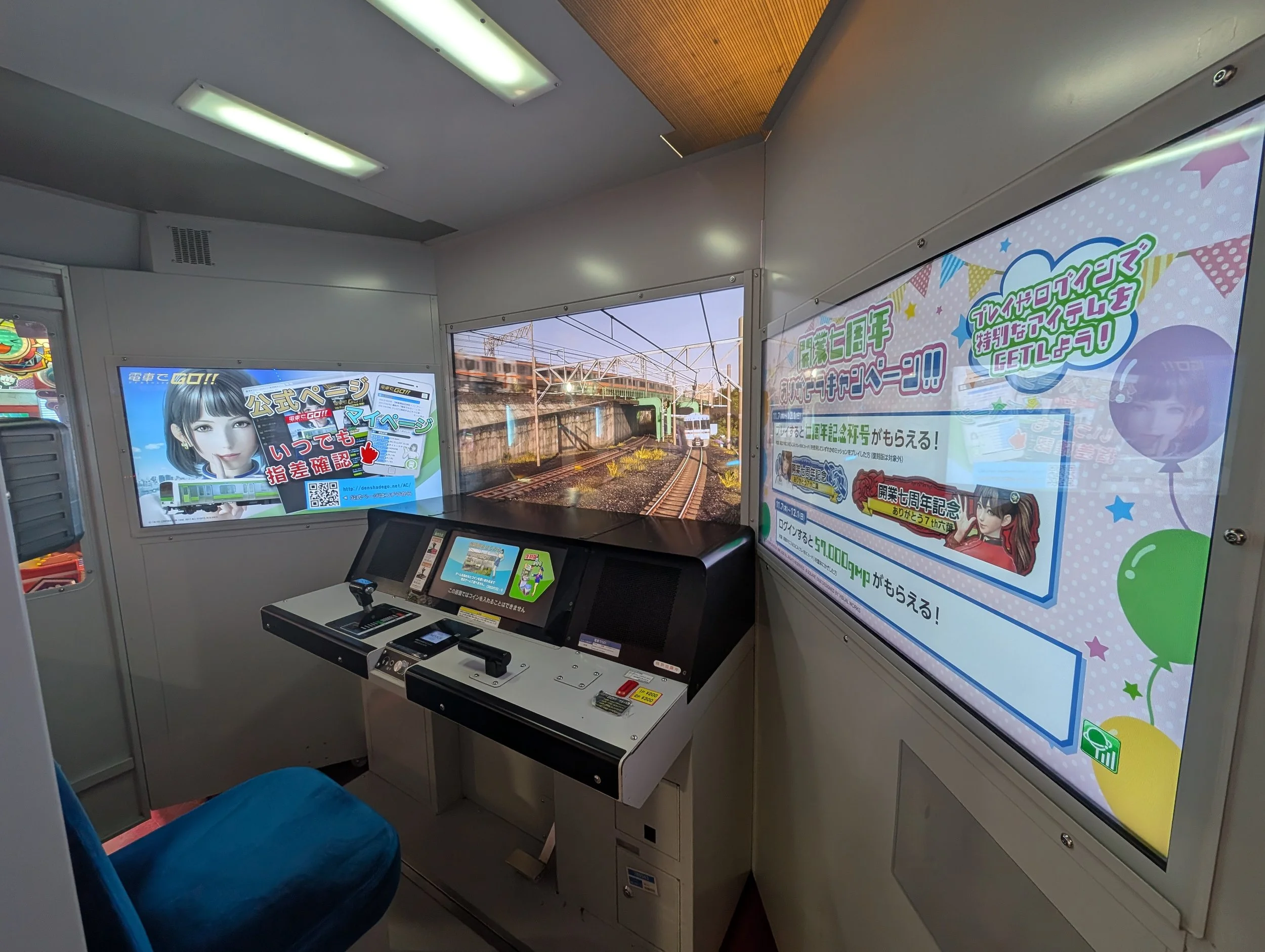 Densha de Go arcade cabinet built as a train driver's cab: throttle and brake controls facing a screen showing the track ahead, with Japanese promotional posters on both walls.
