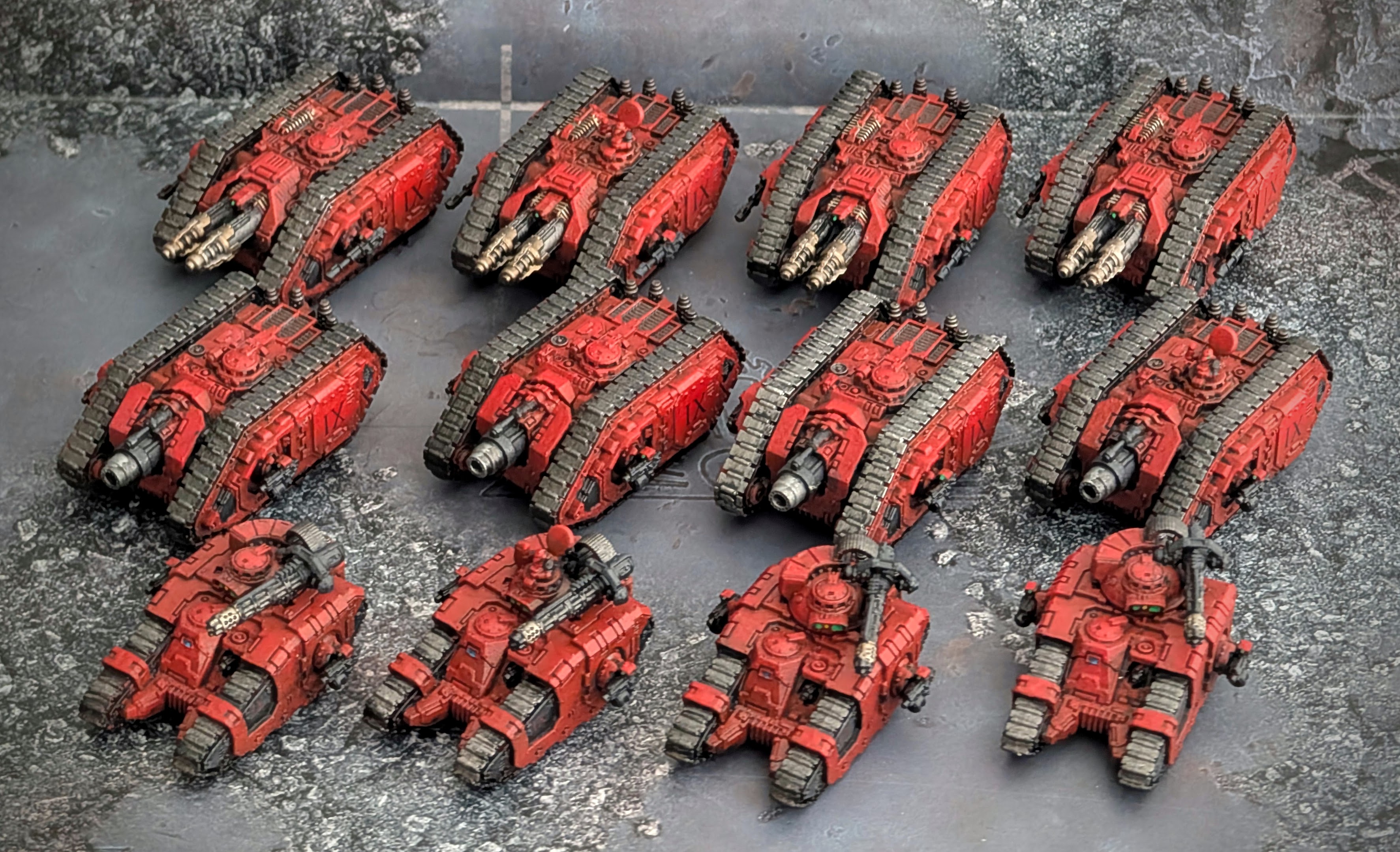 Twelve painted red Blood Angels tanks arranged in three rows on a grey surface, heavier hulls at the back and lighter ones in front.