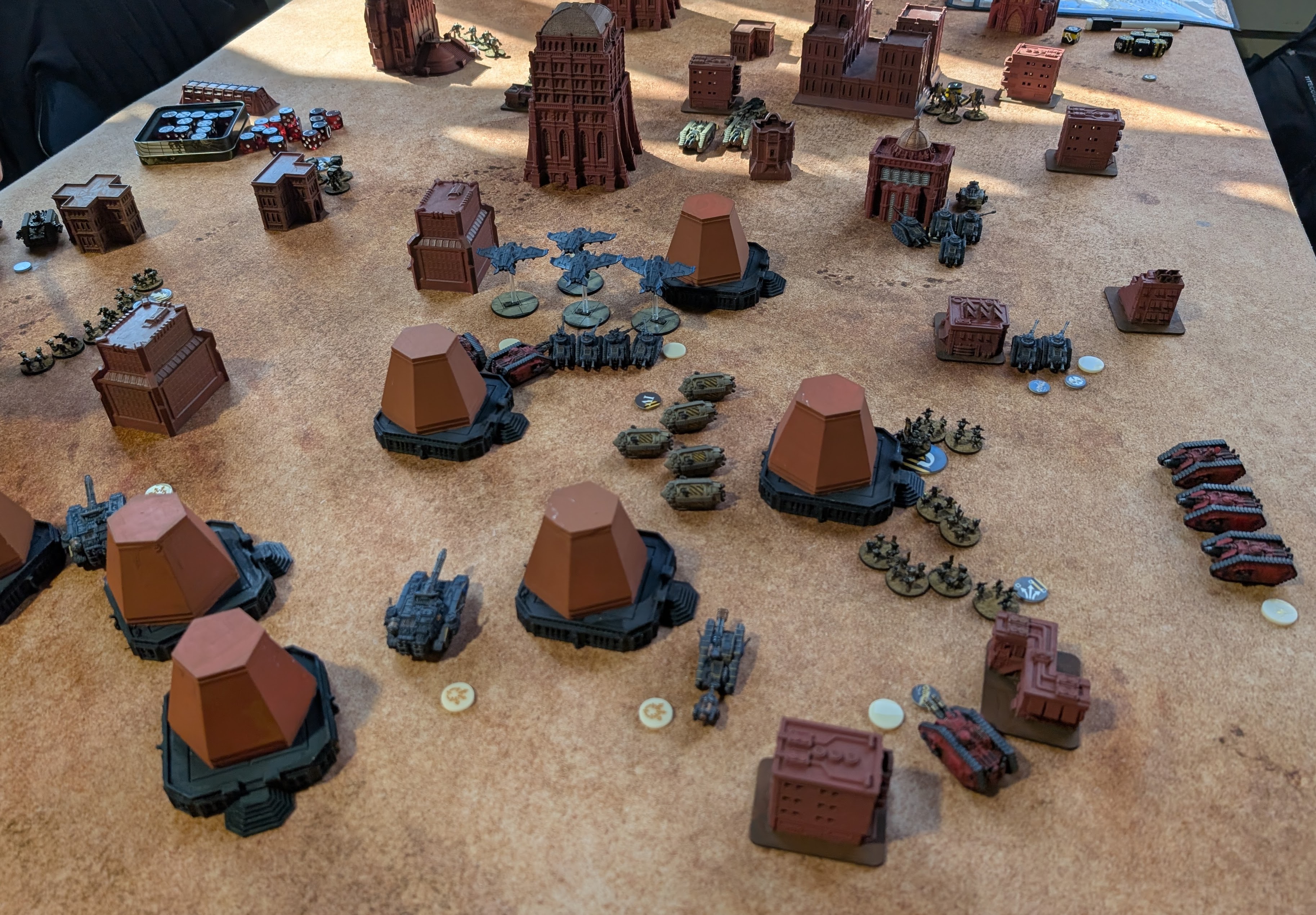 Legions Imperialis tournament table under sunlight: red-primed buildings and terracotta bunkers with grey and red tank squadrons and a flight of aircraft between them.