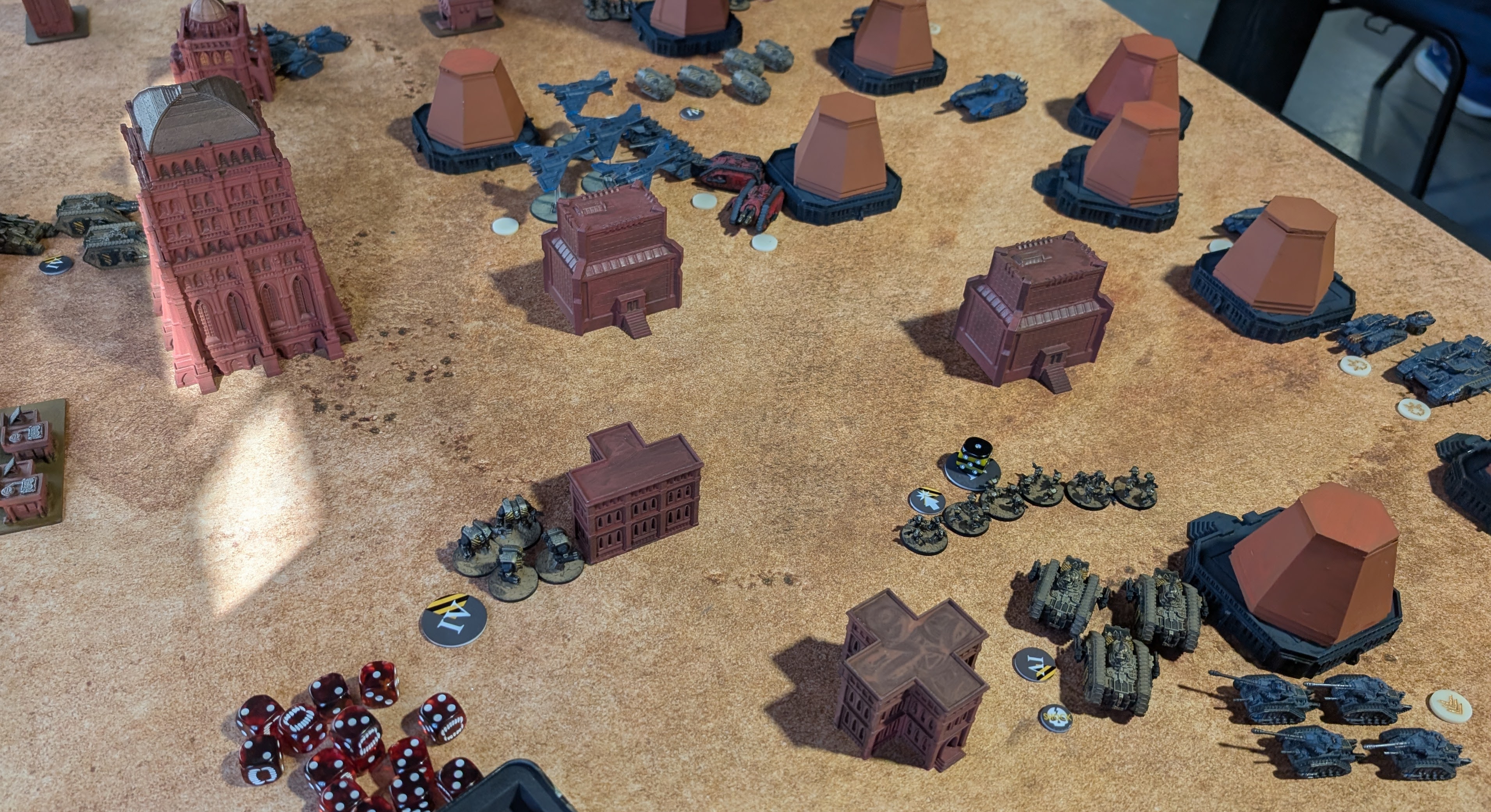 Tournament table with unpainted red-primed buildings on a sandy mat: Iron Warriors infantry and tanks in the foreground, order tokens and a pile of red dice at the table edge.