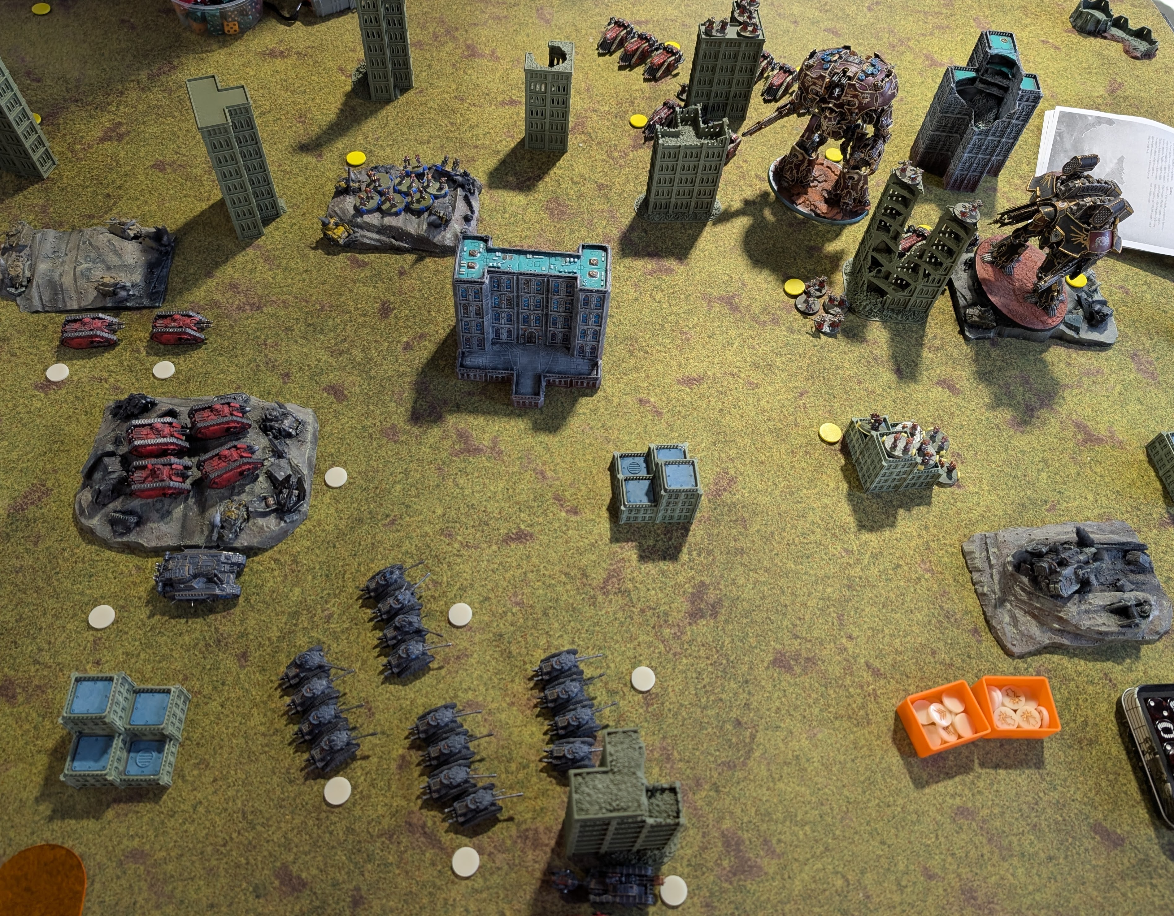 Overhead view of a city table: massed grey tank squadrons and red tanks on hills at the bottom, a towering red-brown Mechanicum titan and a black knight among the buildings at the top.