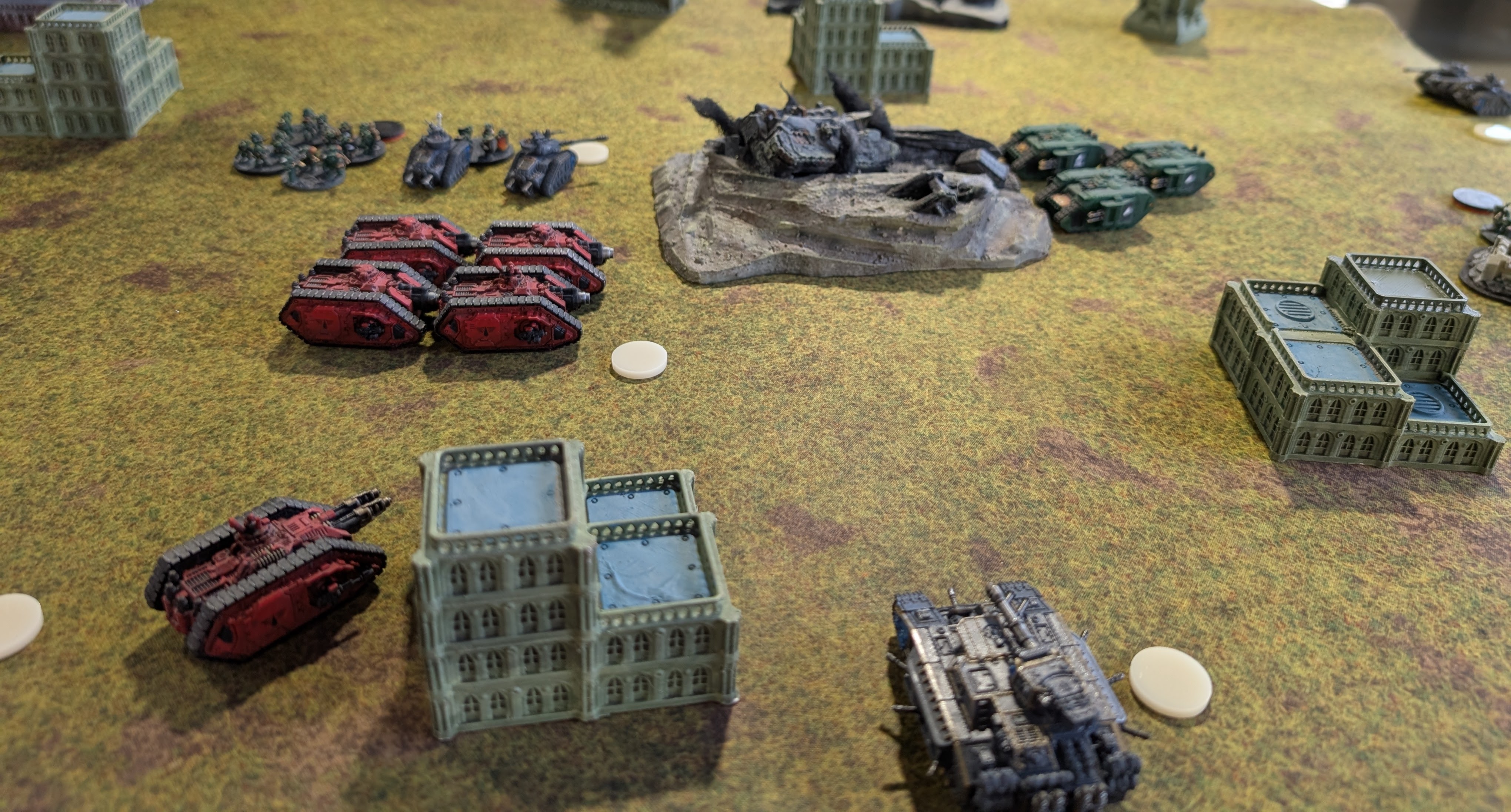 Red Blood Angels tanks facing green Salamanders vehicles and grey Solar Auxilia across a yellow-green mat, a wrecked tank on a rocky outcrop between them.