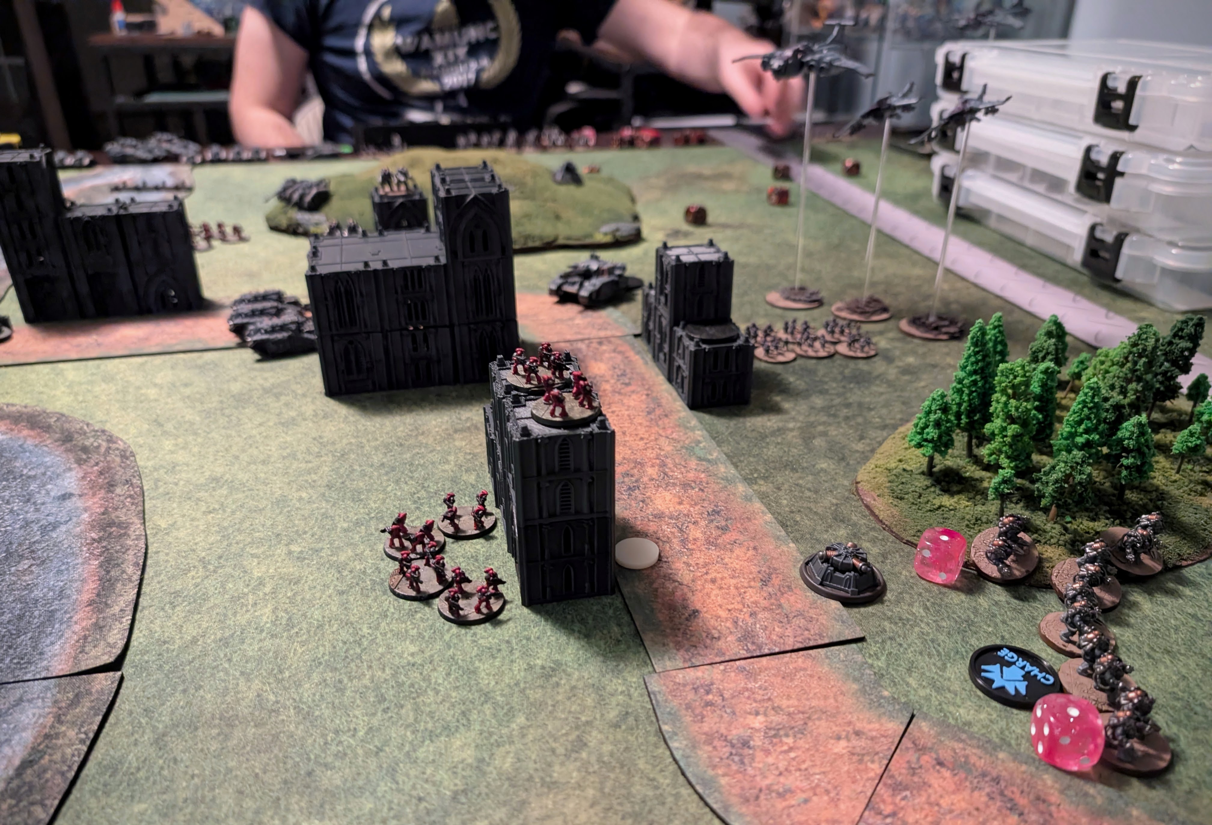 Legions Imperialis game in progress: red-armoured infantry stands garrison black gothic ruins beside a road, with aircraft on flight stands and a charge token on the far side.
