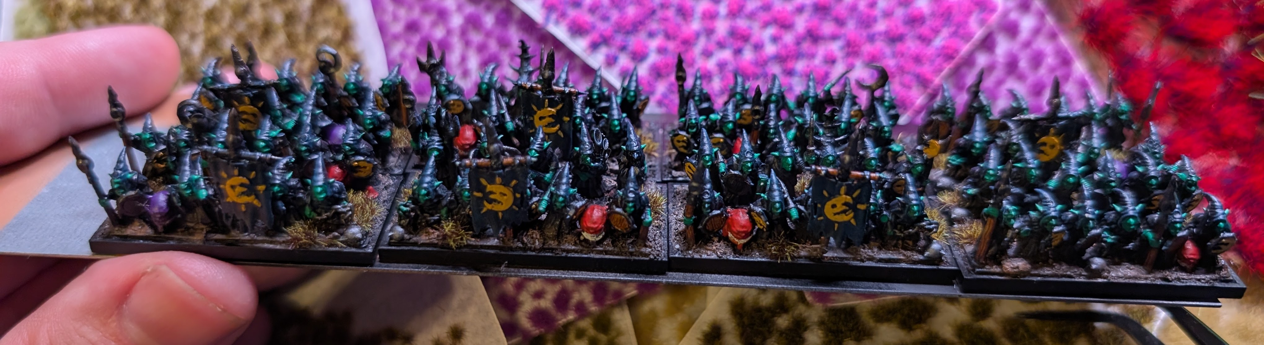 A hand holding a movement tray of painted 10mm Night Goblins in black robes with yellow moon-marked shields.